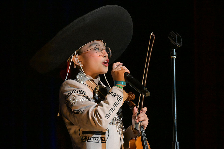 A young girl with glasses holds a violin as she sings into a mic