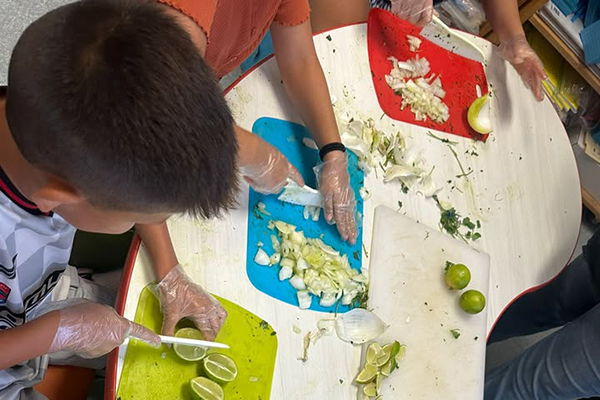 A boy, foreground, cuts a lime on a green cutting board while another boy, background, chops onions on a blue cutting board