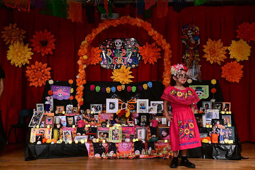 A girl in a pink embroidered dress and flower headpiece stands in front of an ofrenda