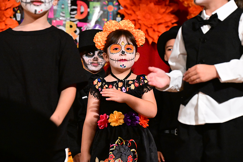 A girl in a black floral dress, orange flower headpiece, and Dia de los Muertos makeup performs on stage