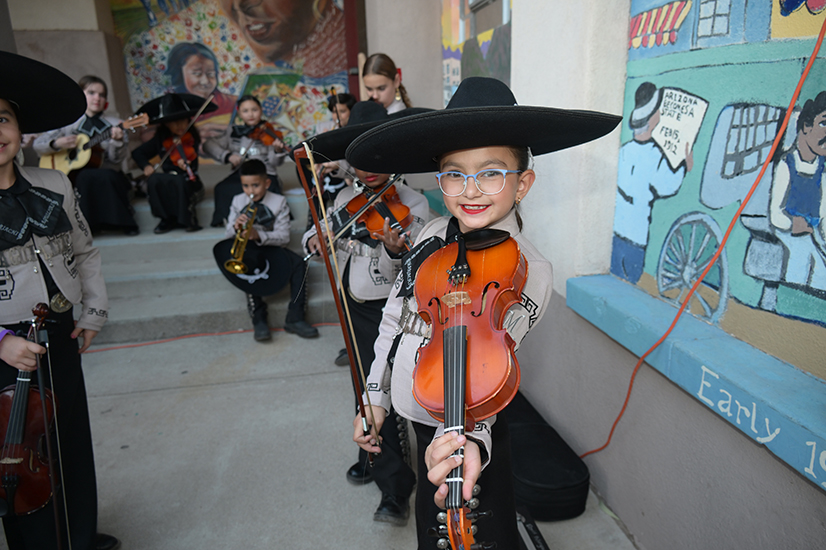 A little girl wearing glasses and a black hat smiles as she holds up her violin