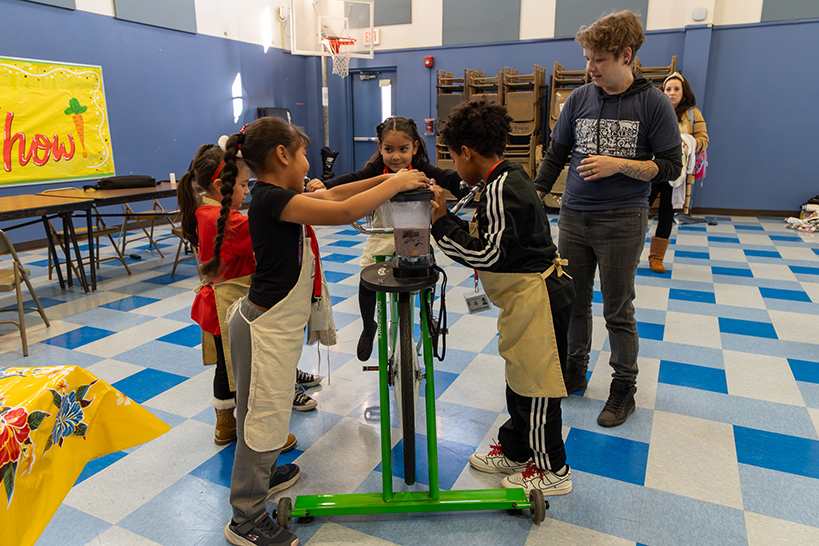 Students gather around as a little girl pedals a bike to blend a smoothie