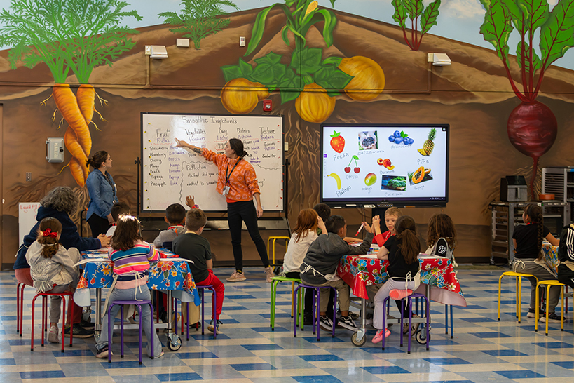 A woman in an orange shirt points to words on a whiteboard, as students sitting around tables listen and watch