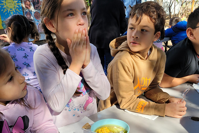 A girl puts her hands to her mouth after eating some soup, while a boy and girl look on