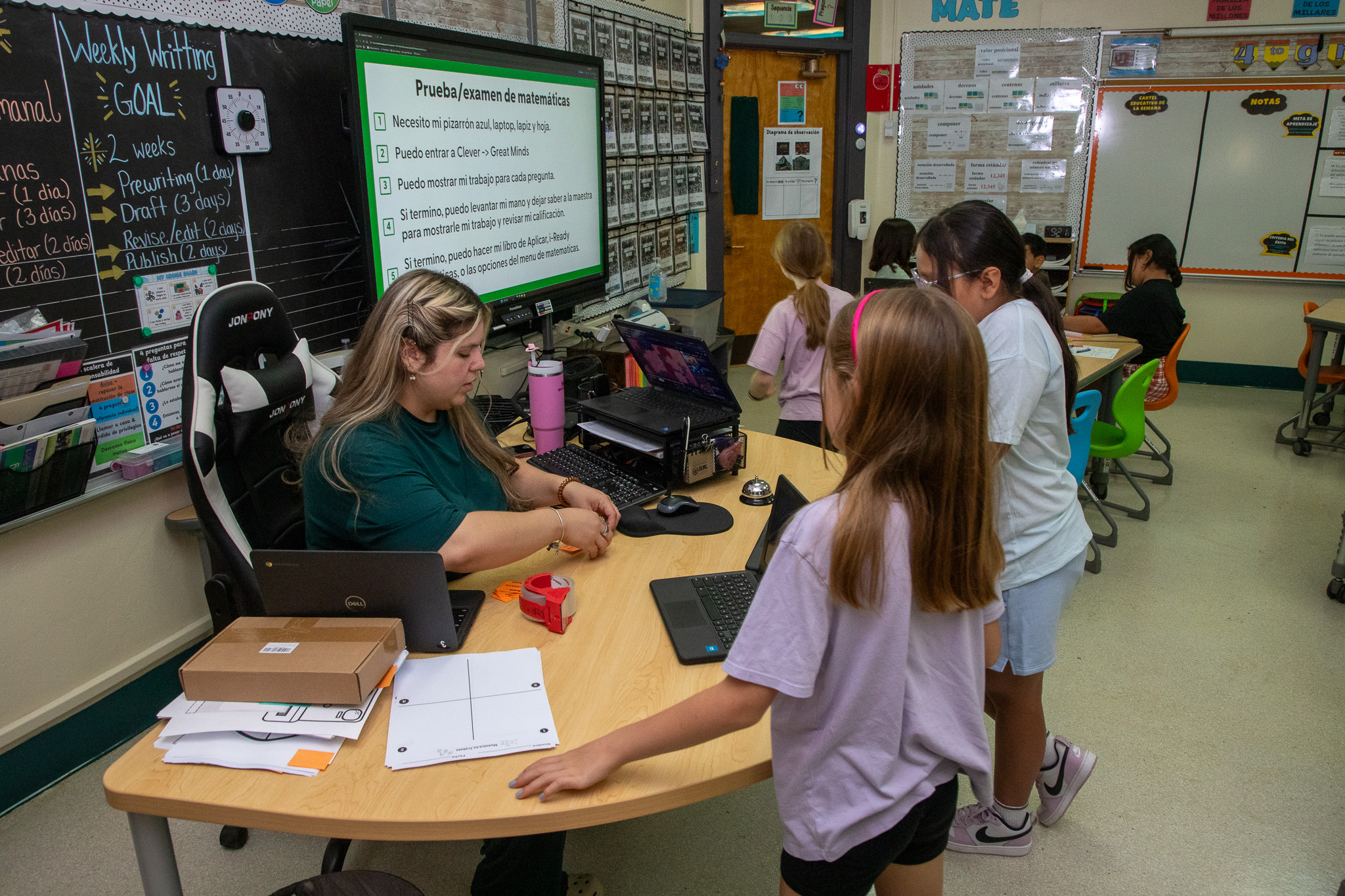 A teacher talks to two of her students as they stand at her desk