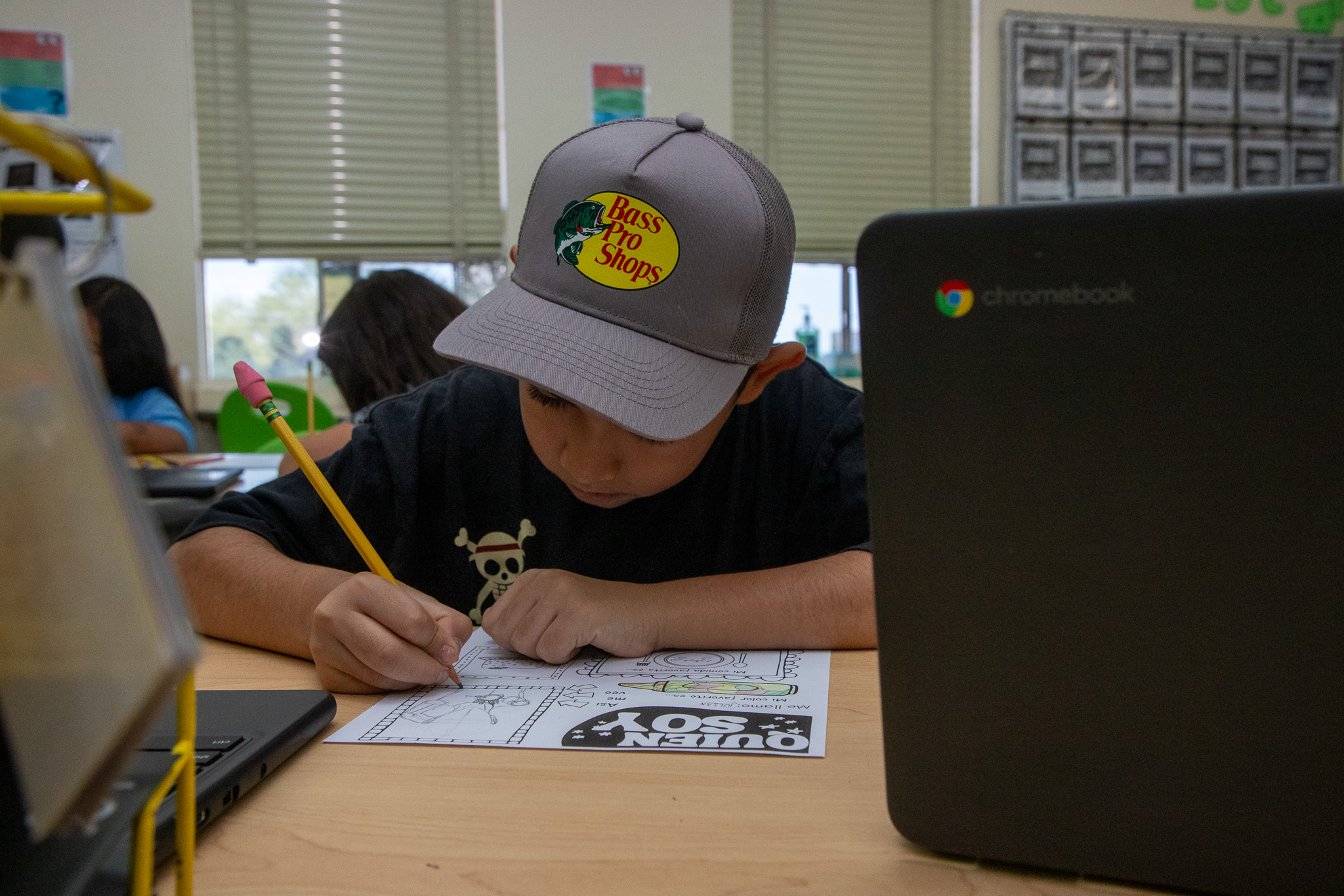 A boy in a Bass Pro Shops hat works on a worksheet at his desk