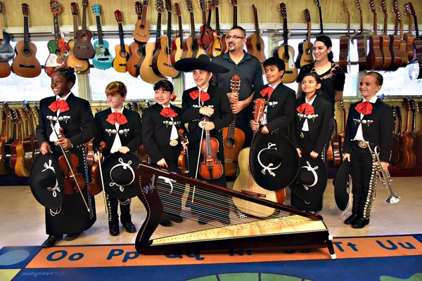 Members of Las Aguilitas De Davis pose in black trajes with program director Jaime Valenzuela. The children are holding instruments and many guitars are hanging on the wall behind them.