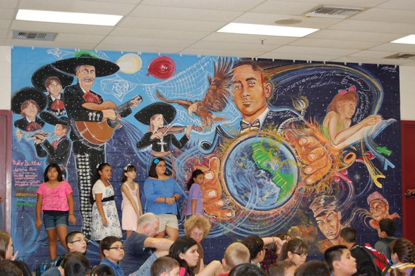Students standing in front of mural of mariachi players, a man holding the world, an eagle, and a child reading.