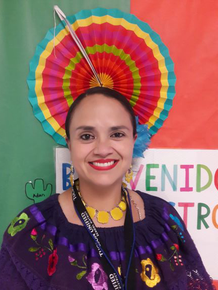 TA Adriana Romo wearing flowery clothing stands in front of a sign reading "Bienvenidos"