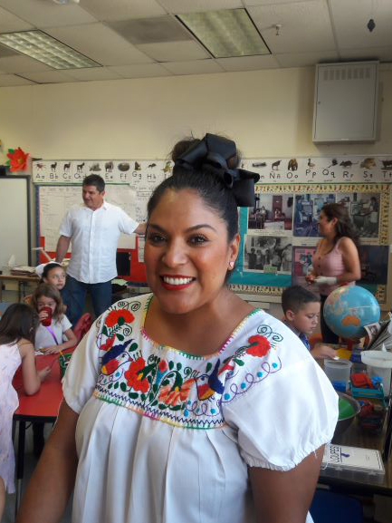 April Cruz smiling in a classroom wearing a flowery white Mexican shirt