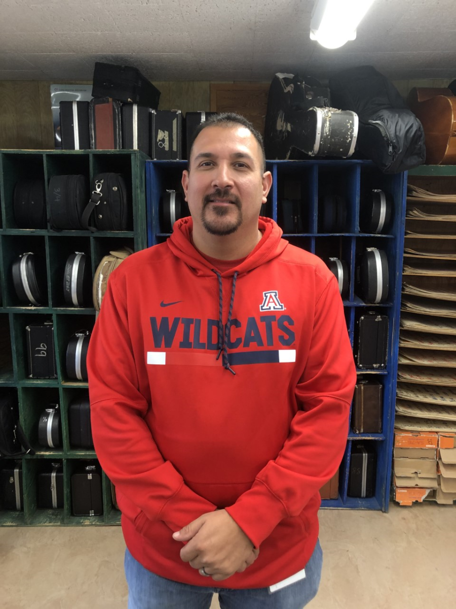 Music teacher Jaime Valenzuela smiles in a Wildcats sweatshirt in front of cubbies filled with instrument cases